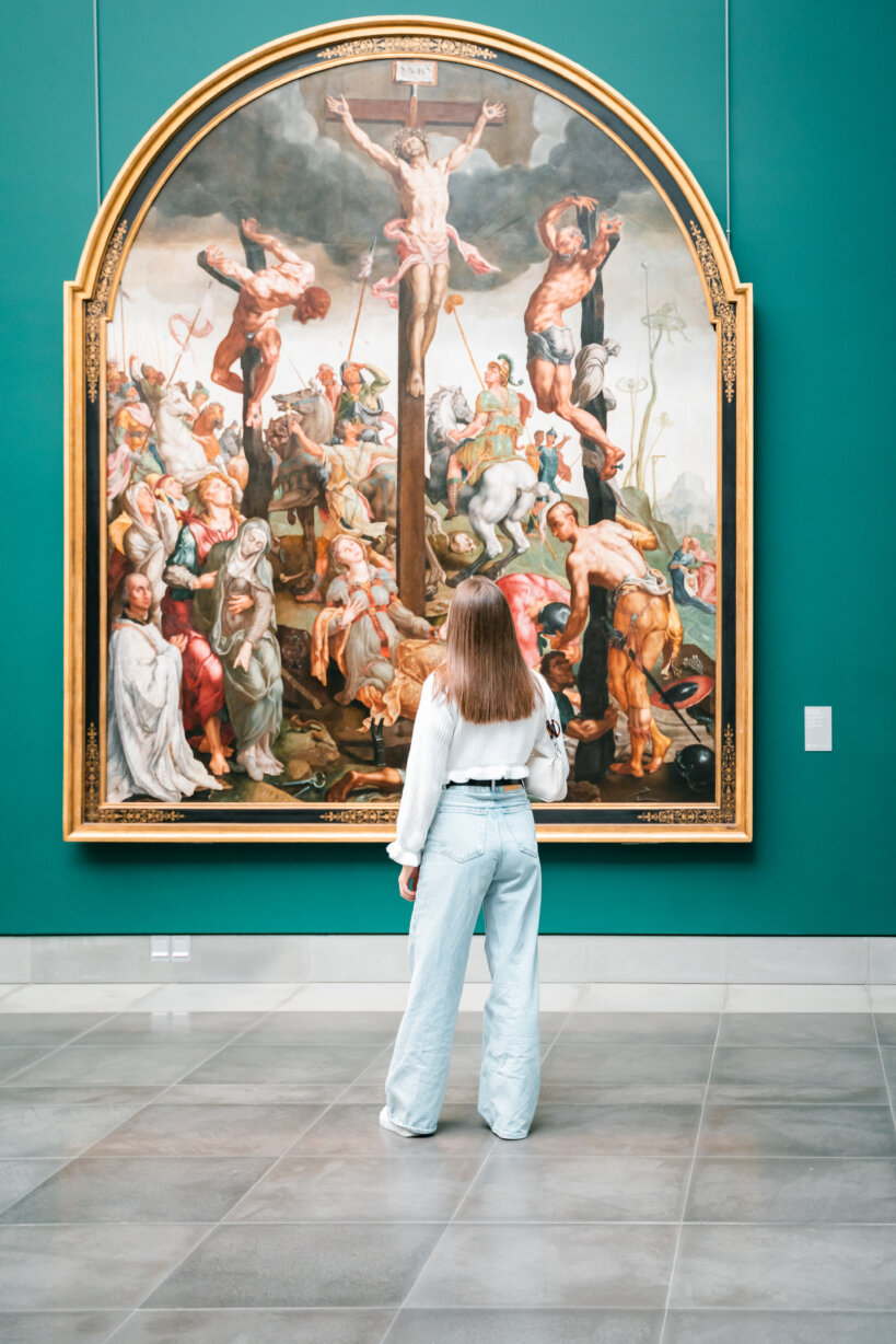 We kijken op de rug van een jongedame met een witte blouse en een lichtblauwe broek. Ze kijkt naar een groot schilderij waarop Jezus centraal staat afgebeeld aan het kruis. Links en rechts van hem, iets meer op de achtergrond, hangen twee andere veroordeelden.