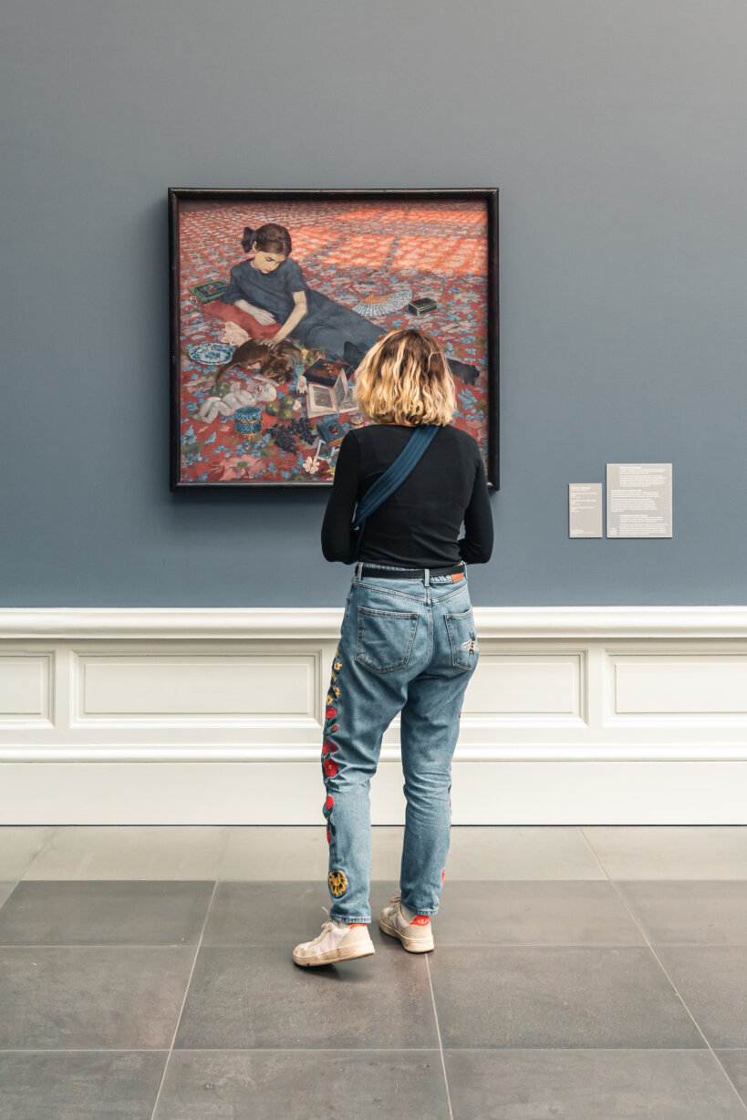 Een vrouw staat voor een schilderij dat tegen een blauwgrijze muur hangt. Op het werk zie je een jong meisje dat op een rood tapijt ligt. Ze draagt een donkerblauw kleedje.