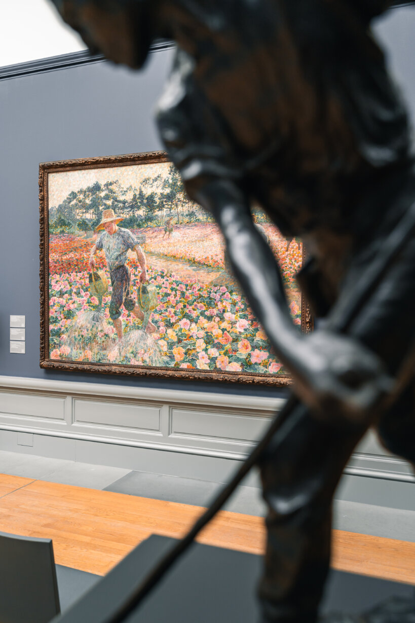 Op de voorgrond staat een zwart standbeeld. Daarachter hangt een werk waarop een tuinier in een veld vol hortensia's is afgebeeld terwijl hij ze water geeft met twee gieters.
