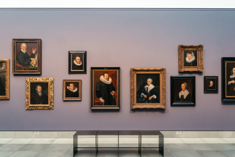 Er hangen 11 portretten aan een grijsblauwe muur. De twee grootste portretten hangen in het midden. Voor de portretten staat er centraal een zitbank.