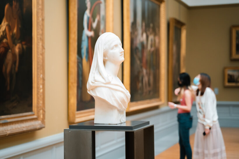 Centraal in beeld staat een witte buste van een vrouw. Daarachter hangen een viertal grote schilderijen uit de achttiende en negentiende eeuw. Er staan op de achtergrond twee vrouwen te kijken.