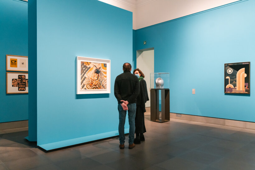 Een man en een vrouw praten met elkaar voor een schilderij dat aan een brede blauwe zuil hangt. Achter de brede zuil hangen nog twee schilderijen tegen een blauwe muur. Aan de rechterkant staat nog een blauwe muur waar een schilderij aan hangt.